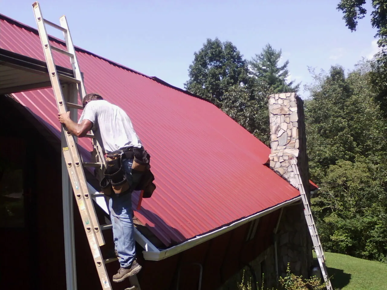 Roofer on a ladder beside a red metal roof and stone chimney — Soffit & Fascia Repair by Roofing Brothers in Funston